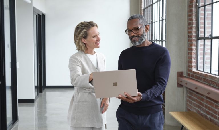 Man and woman standing in an office hallway discussing content on a laptop.