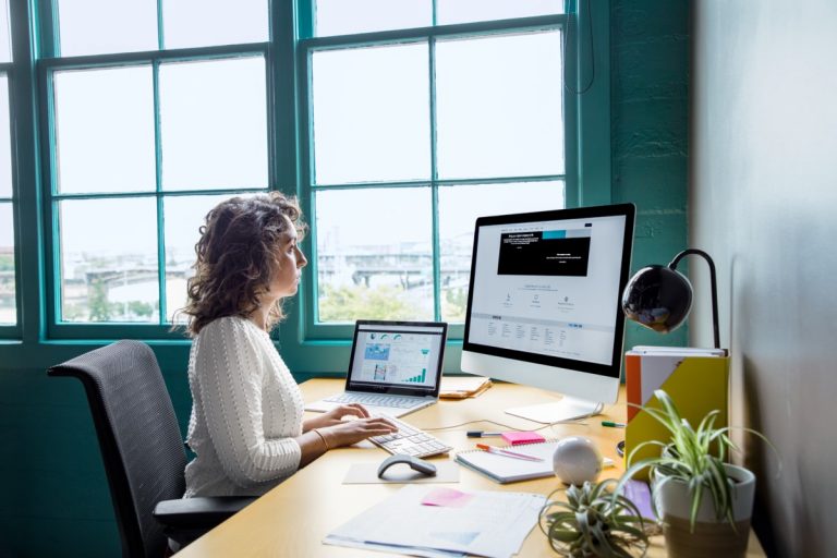 Female office worker sitting at desk using an all-in-one computer in casual office with a laptop also on desk.