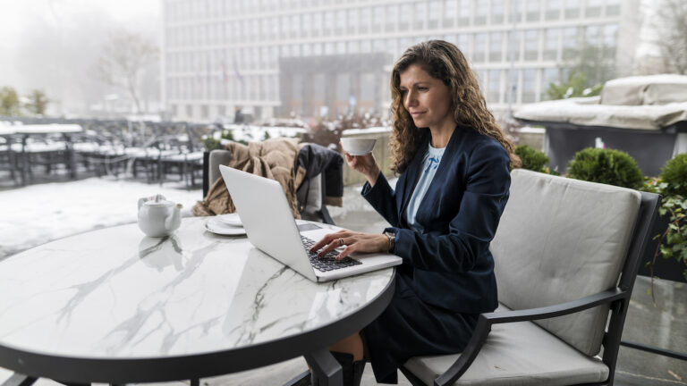 Businesswoman in a blue jacket, sitting at a table outdoors during the winter, checking her laptop while drinking coffee out of a white mug / sandbox preview