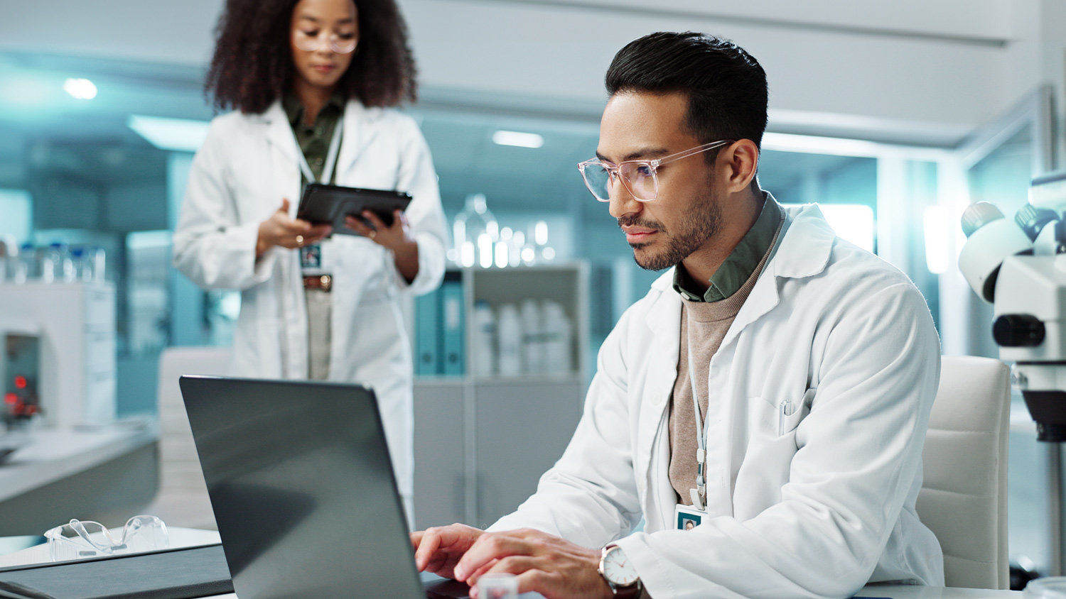 AI for life sciences helps two pharma techs wearing lab coats review trial information on tablet devices.