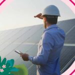 A mobile worker stands next to solar panels, shielding his eyes while looking toward the sun and a bright future.